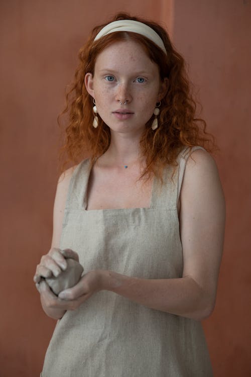 Peaceful woman with red hair and freckles wearing apron and looking at camera while standing near wall with piece of clay in hands