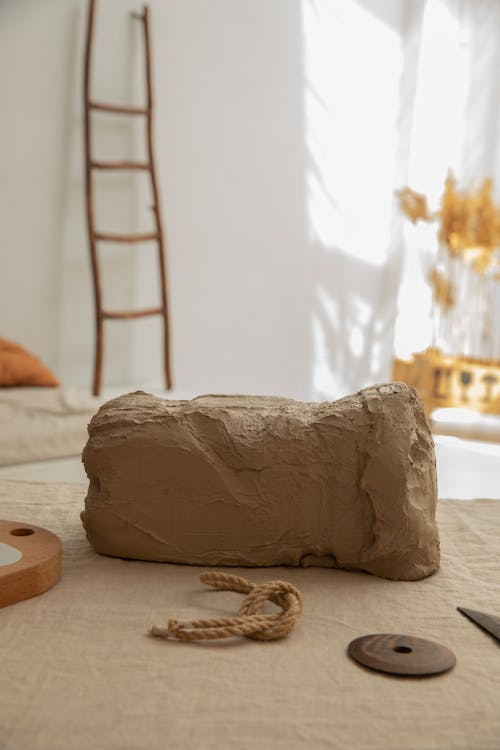 Chunk of natural clay placed on workbench with textile in light room with wooden ladder and plants on blurred background