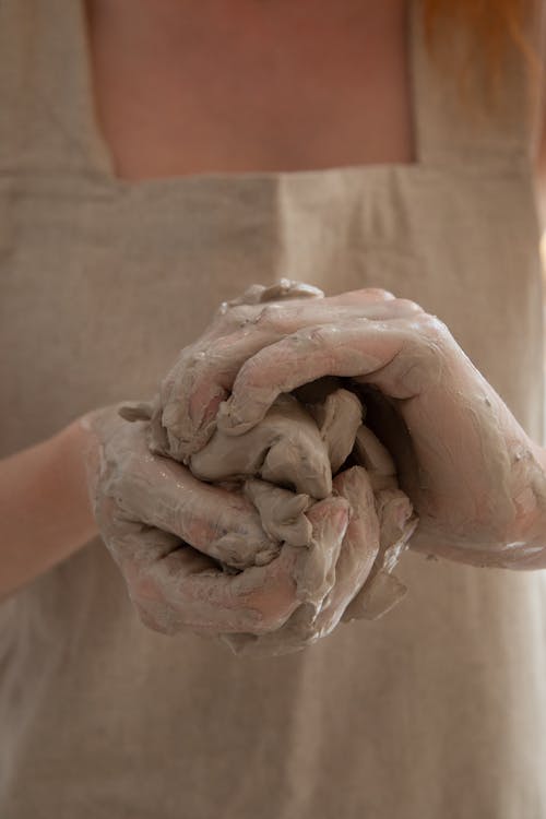 Free Unrecognizable person wearing gray apron shaping and molding clay in dirty hands while making handicraft crockery in workshop during work Stock Photo
