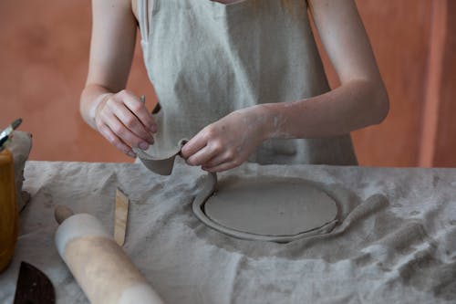 Unrecognizable skilled female master in apron standing near table with rolling pin and working with clay while making crockery during work in pottery workshop with blurred background