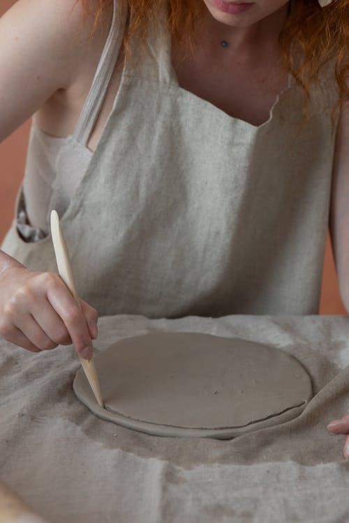 Unrecognizable female master in apron cutting round piece of clay with knife while making pottery during work in professional studio