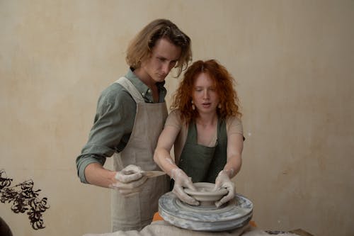 Focused couple in aprons with dirty hands modelling clay products on wheel while working together at home