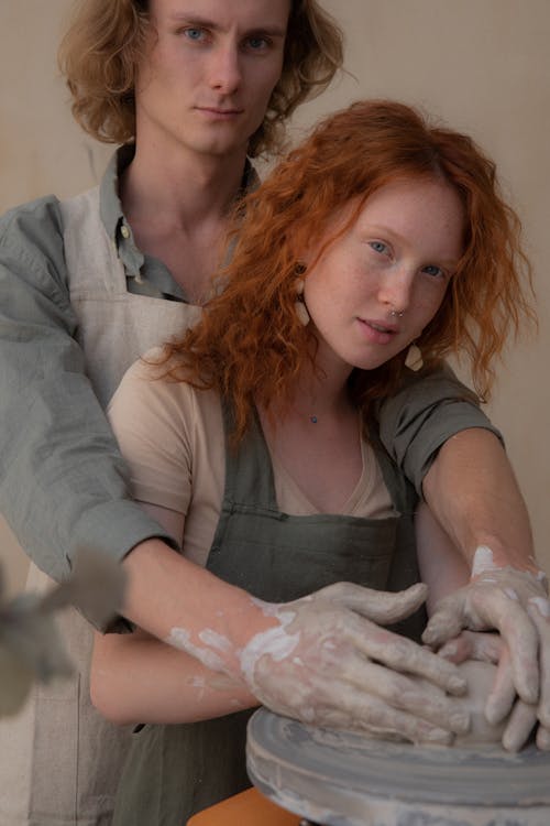 Crop young couple in aprons hugging while modelling clay products together and looking at camera