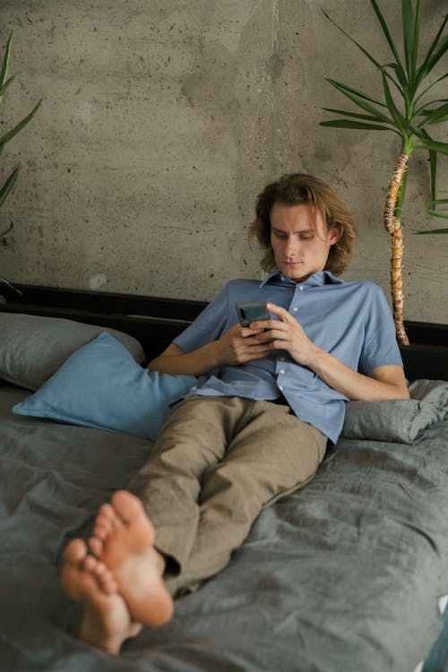 Free Relaxed barefoot male in casual wear lying on comfortable bed while messaging on mobile phone and looking at screen Stock Photo