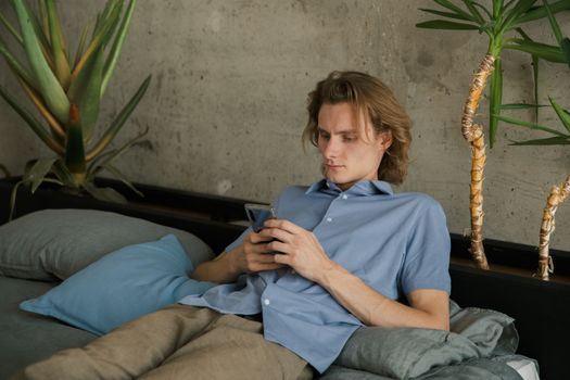 一个穿着休闲服的年轻男子躺在床上，正用智能手机看着屏幕
