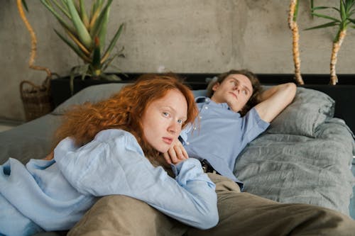 Free Ginger haired female looking at camera and leaning on leg of boyfriend while lying on bed during weekend in room with plants Stock Photo