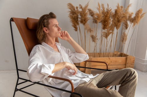 Side view of pensive male in white shirt sitting on comfortable armchair with hand at chin and looking in distance in room with dried plants