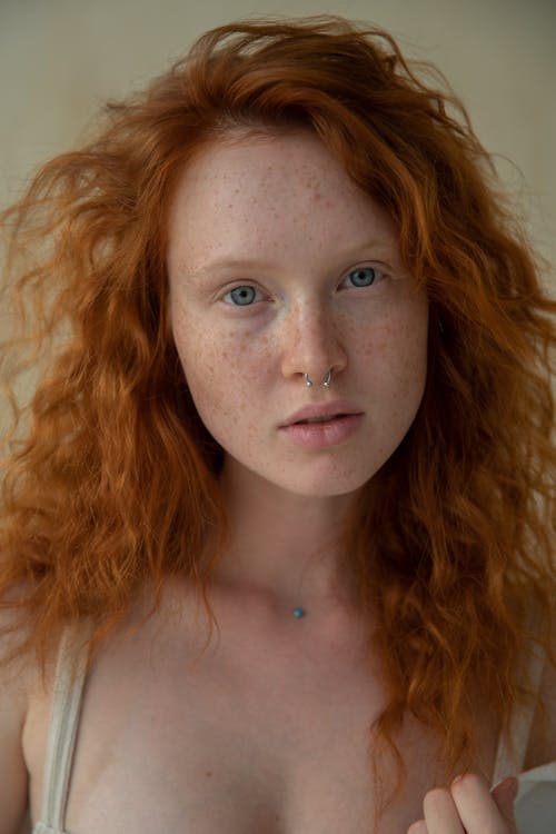 Portrait of dreamy female with curly red hair and freckles with pierced nose looking at camera while standing on background