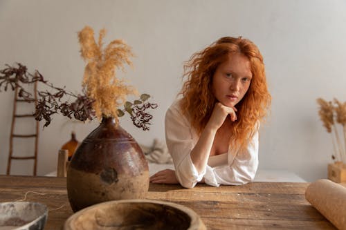 Dreamy female with red hair looking at camera while leaning on hand at table with dry flowers in vintage vase