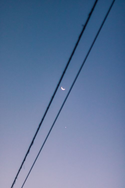 Free Crescent moon visible behind electricity wires against a twilight sky, creating a serene scene. Stock Photo