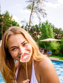 A cheerful woman holding a lipstick cutout by a swimming pool.