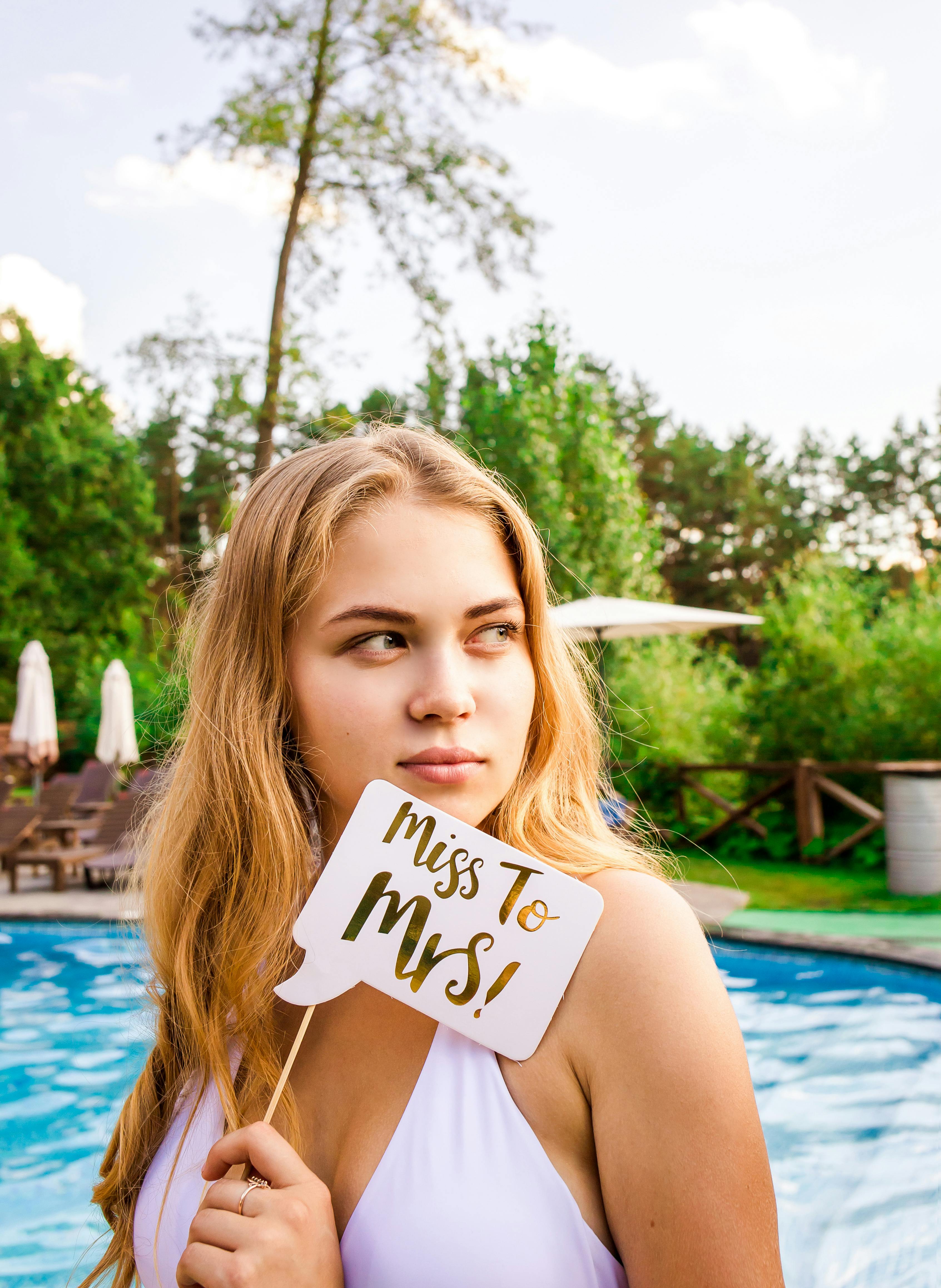 Woman in bikini beside pool holding "Miss to Mrs!" sign, perfect for bachelorette themes.