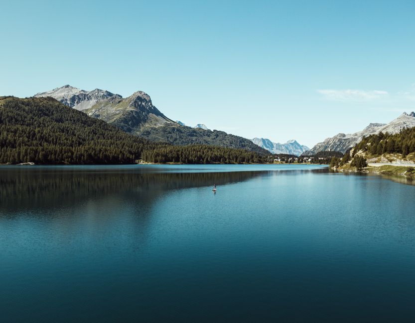 Peaceful mountain lake surrounded by lush forests and distant peaks under a clear blue sky.