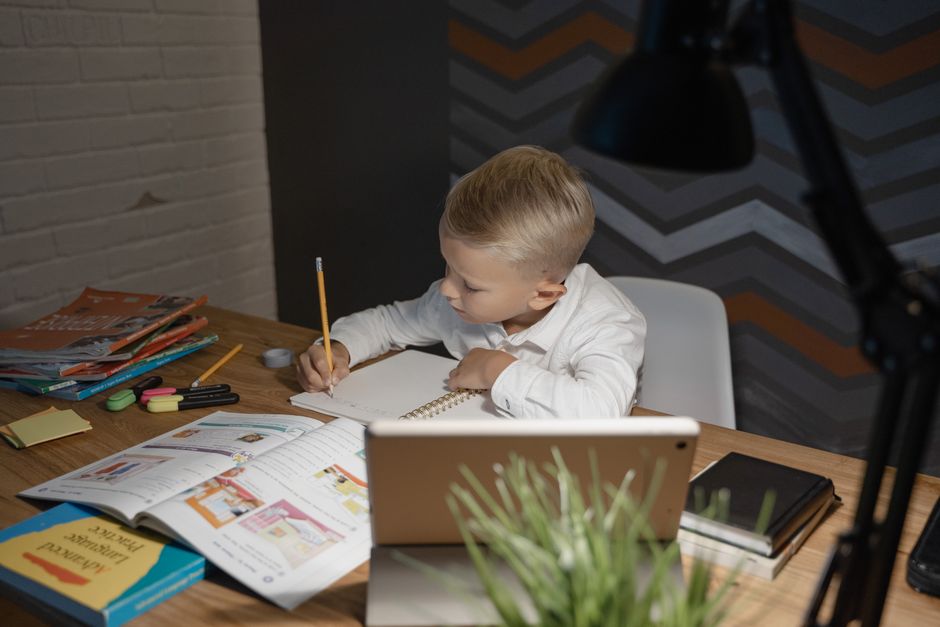 A young boy focuses on homework with books and a tablet at a desk, enhancing his education.