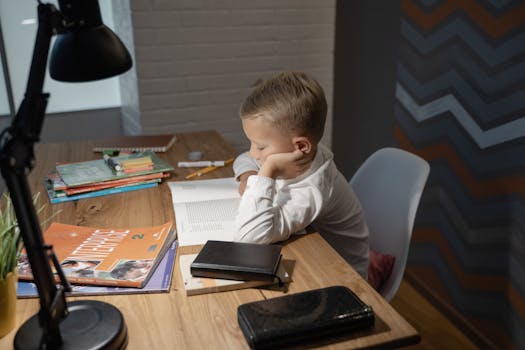 A young boy is reading a book at a desk with study materials and a desk lamp indoors.