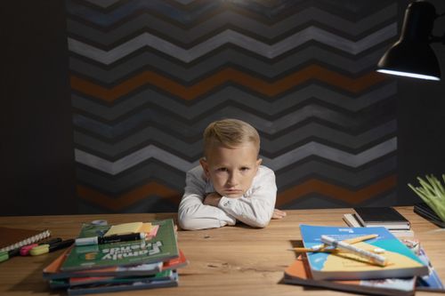 Free A young boy leaning on a table surrounded by books and coloring materials, looking unhappy with studying. Stock Photo