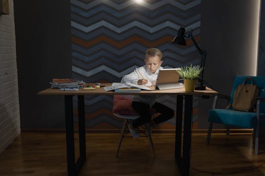 A young boy focused on his homework at a desk with a tablet, lamp, and books.