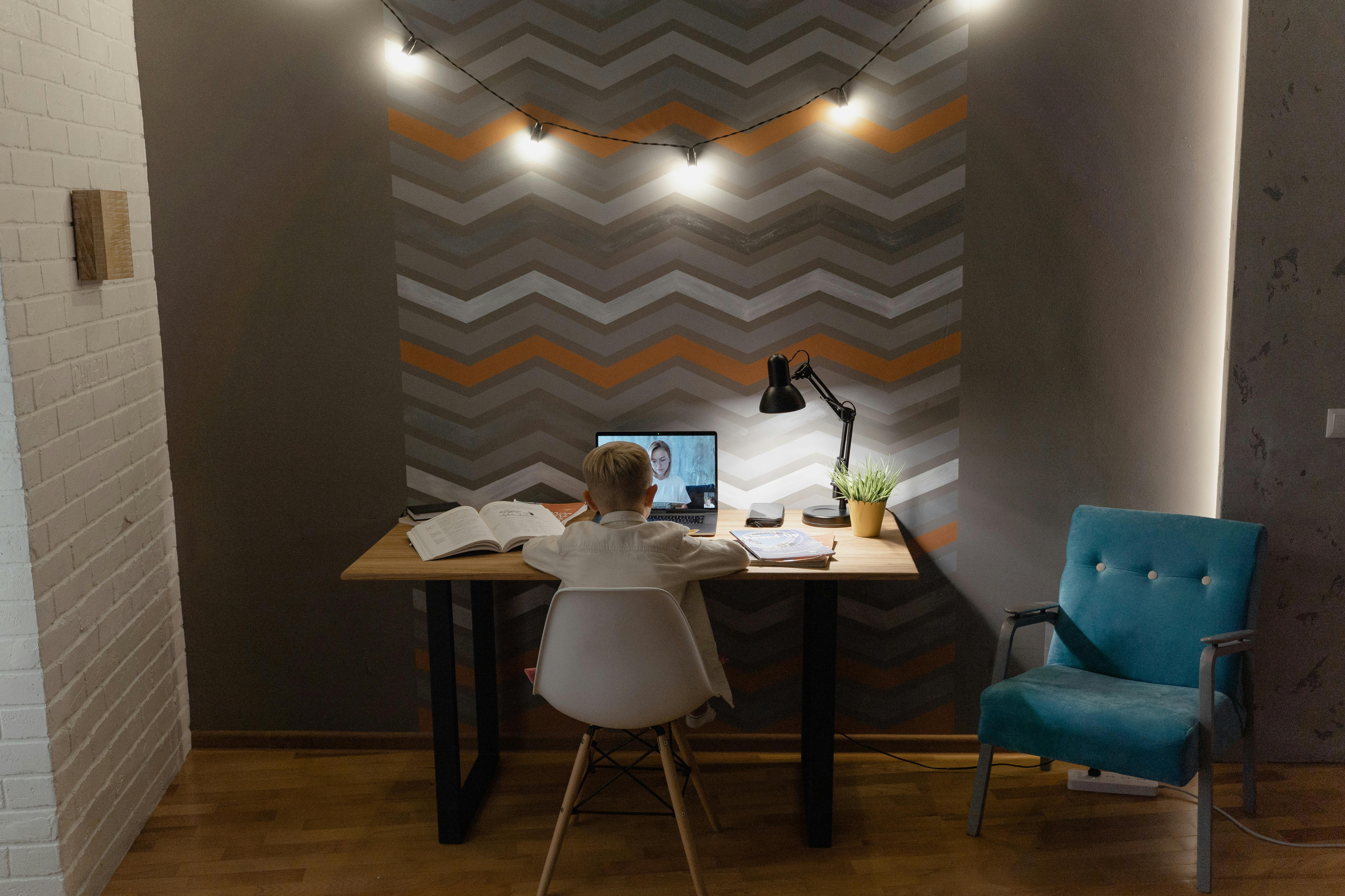 一个小男孩在课桌上参加在线课程，桌上放着书和电脑