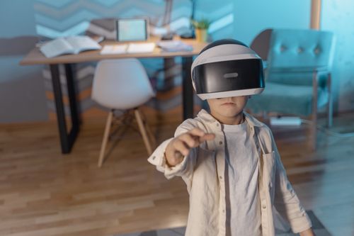 Free Young boy wears VR goggles, exploring virtual reality indoors with books open on desk nearby. Stock Photo