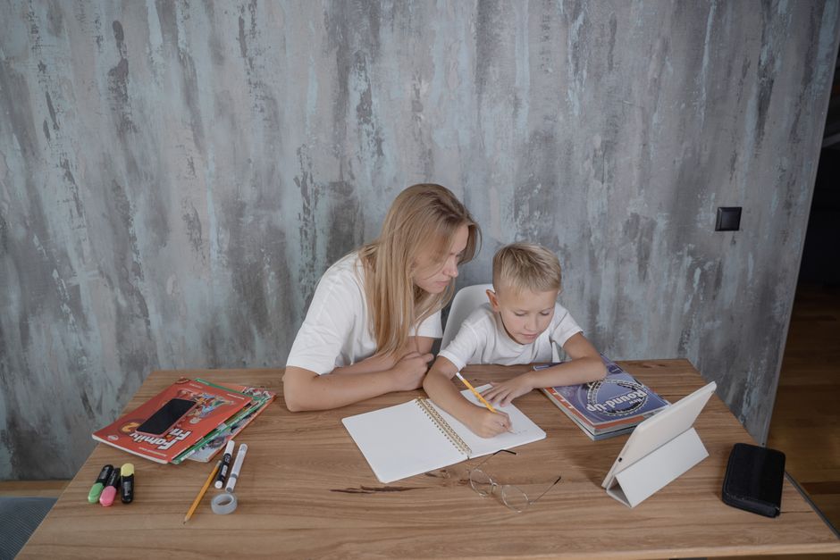 A mother guides her young son during a homework session at a desk in a cozy home setting.