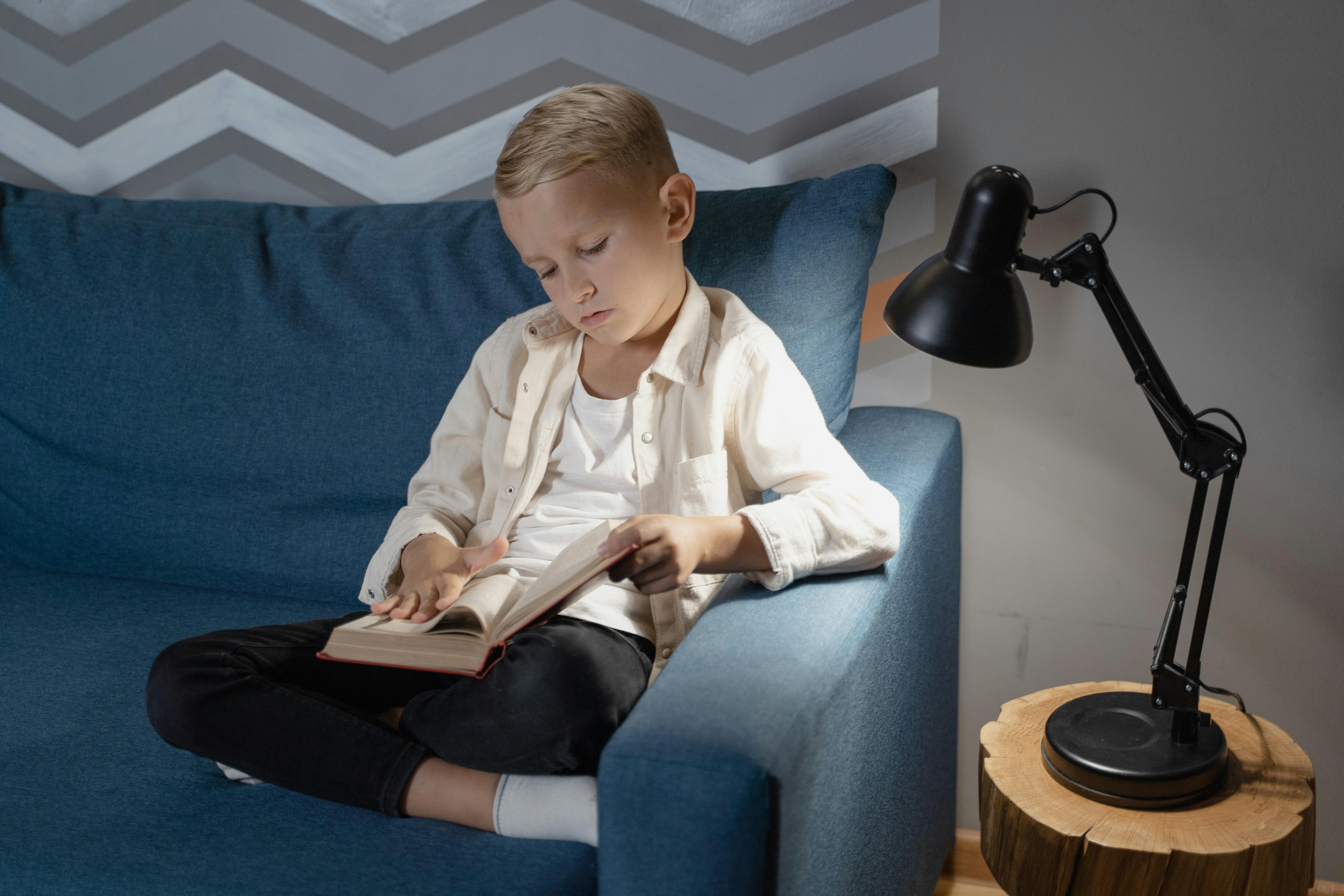 Free A young boy attentively reading a book while sitting on a cozy blue couch indoors next to a lamp. Stock Photo