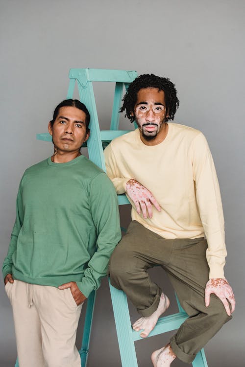 Confident multiracial male models in trendy outfits sitting on ladder against gray background and looking at camera