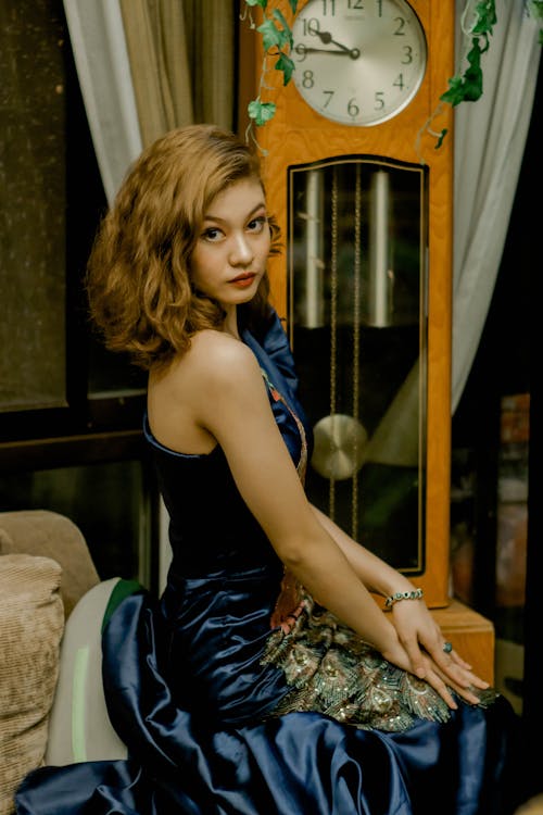 Side view of gorgeous female wearing stylish dress sitting near vintage clock and looking at camera