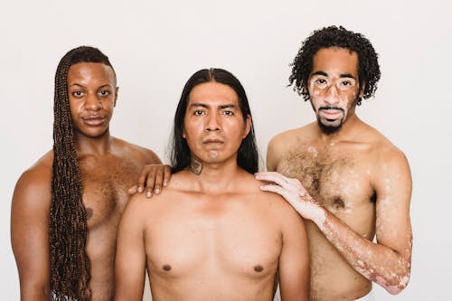 Serious multiracial male models holding hands on shoulders and looking at camera while standing against white background