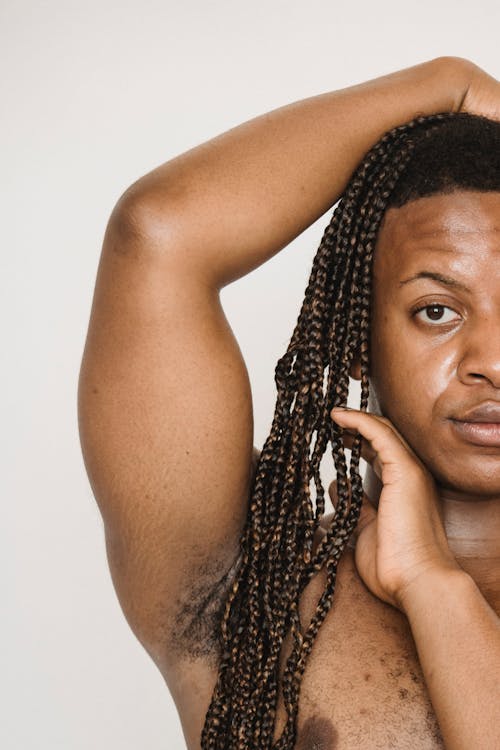 African American transgender with naked torso touching Afro braids and looking at camera while standing with hand on head on white background