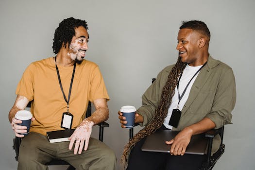 Smiling African American male in casual wear with vitiligo skin sitting on chair and talking to androgynous male coworker with laptop during coffee break