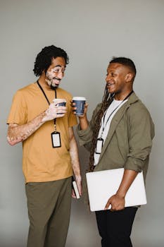 Cheerful African American male living with psoriasis wearing casual clothes talking to content black coworker while chatting and drinking hot takeaway coffee together