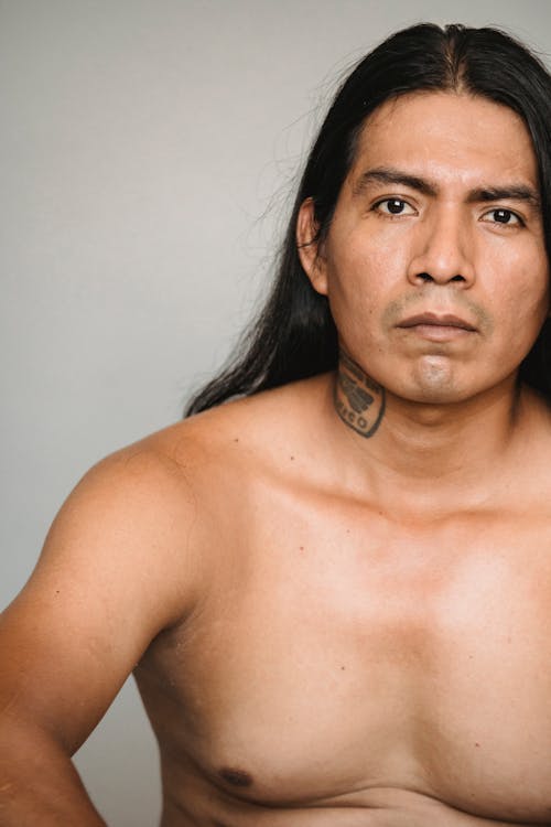 Pensive American Indian male with long dark hair and naked torso looking at camera against gray background in studio