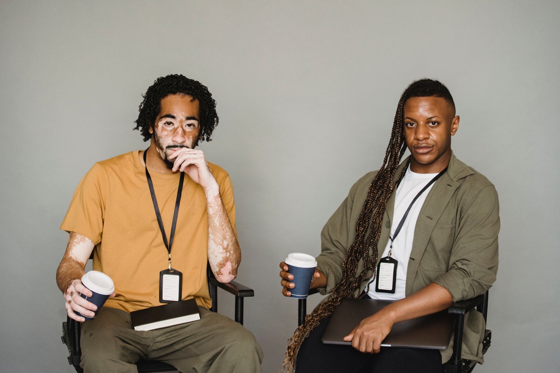 Serious African American colleagues in casual clothes with name tags and cups of takeaway coffee sitting in studio and looking at camera against gray background