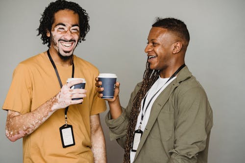 Cheerful black man with vitiligo skin laughing and drinking coffee with androgynous black man with afro braids against gray background