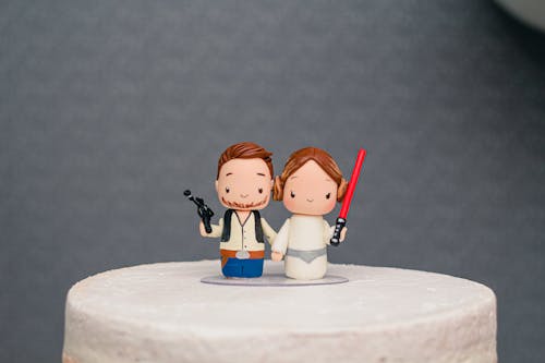 Funny figures of couple holding hands decorating delicious wedding cake placed against gray background