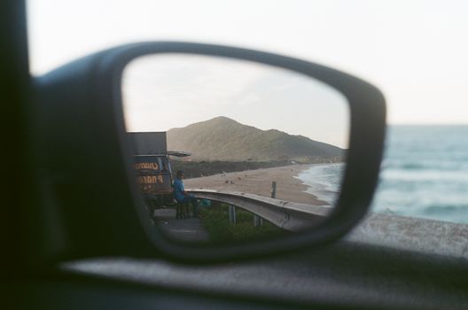 View of Florianópolis beach in car side mirror with mountain backdrop, capturing serene coastal landscape.