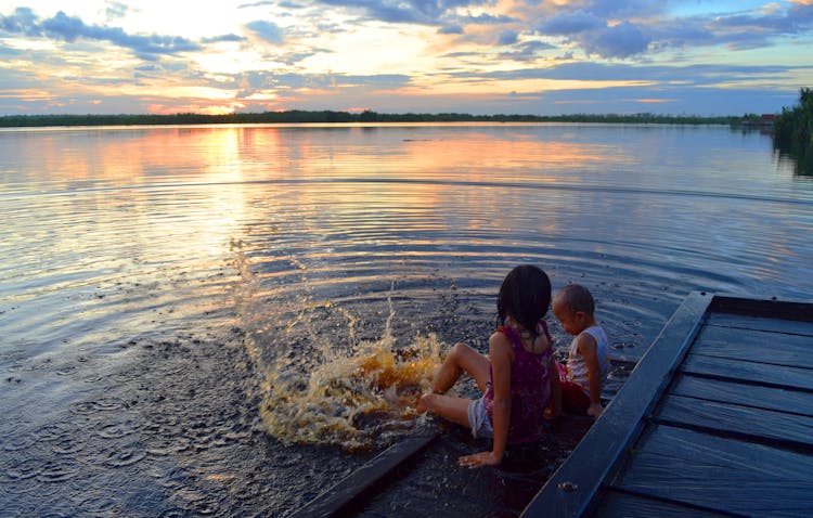 Children Sitting On Wooden Dock By The Lake