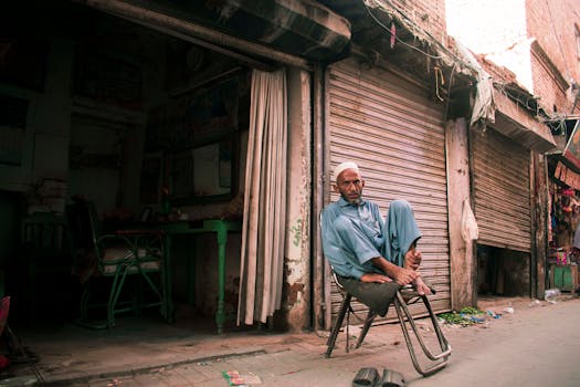 Elderly man in traditional wear sitting in front of a shop with roll-up doors on a street.