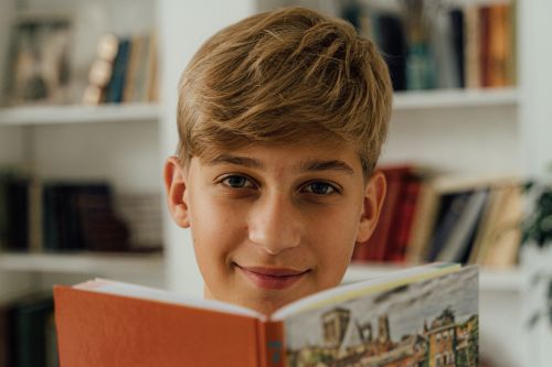Free Blond young student smiling and reading a book in a library setting, promoting education and happiness. Stock Photo