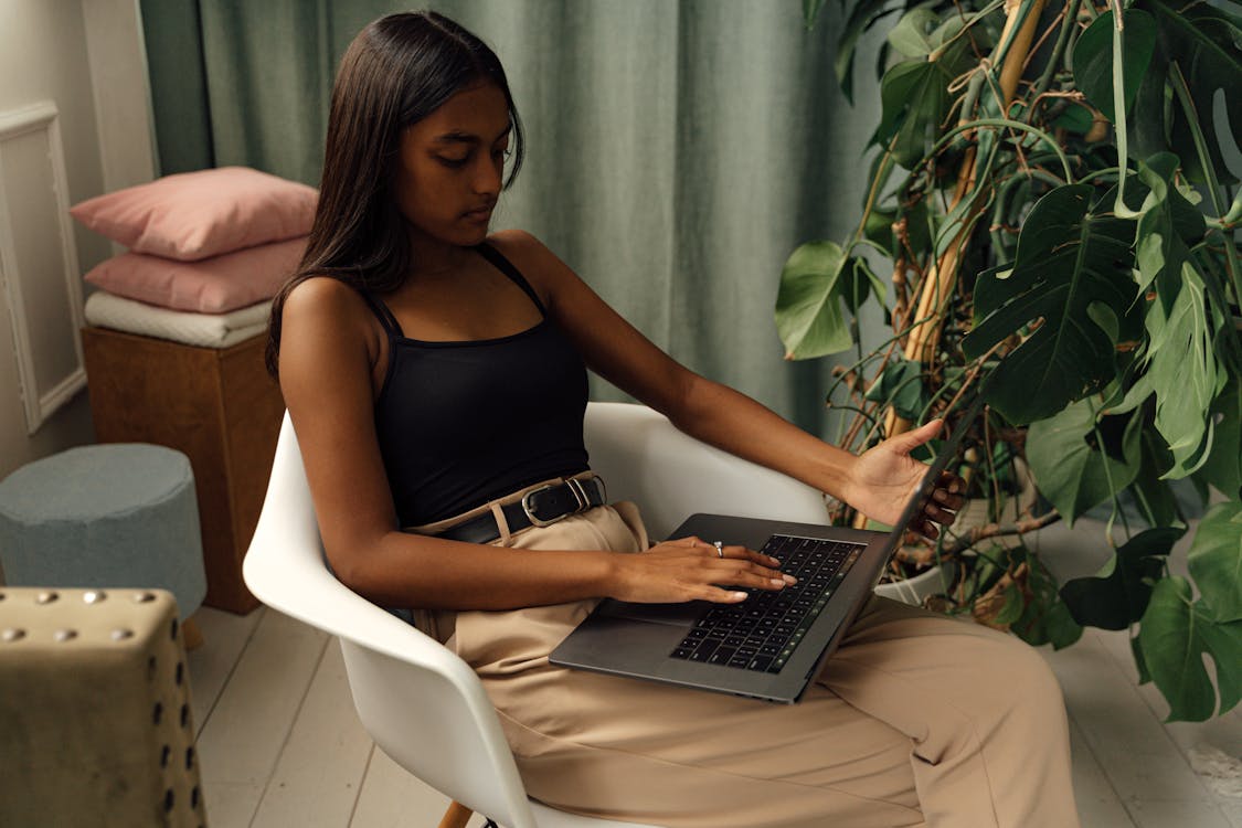 Free A young girl sits in a chair using a laptop, surrounded by plants in a cozy indoor setting. Stock Photo