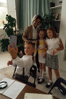 Group of children happily taking a selfie with their teacher in a bright classroom setting.