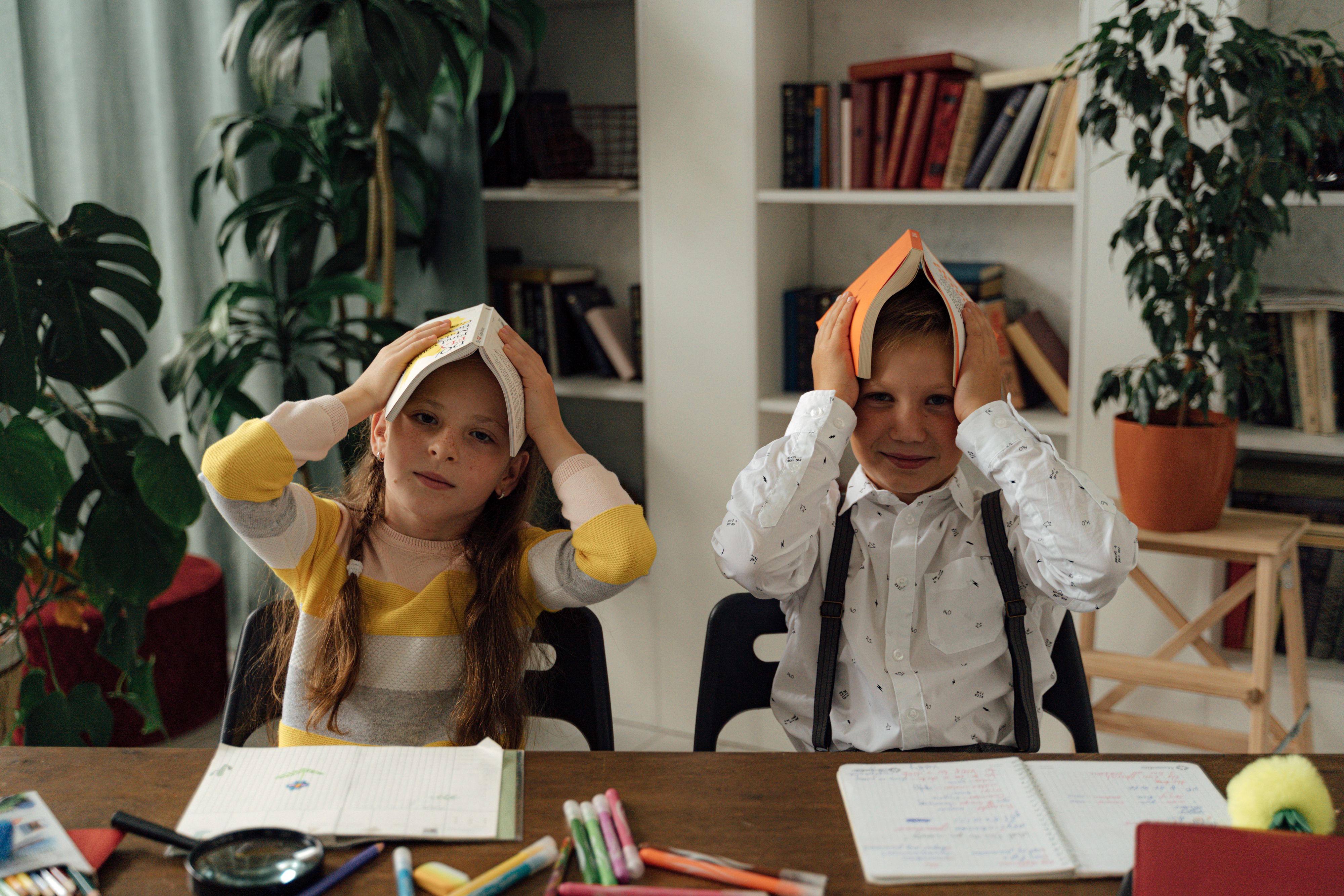 Two children playfully place books on their heads during a study session indoors.