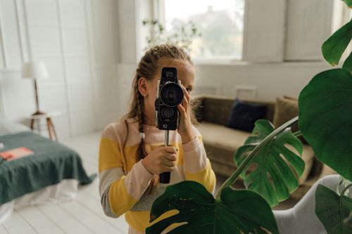 Free A young girl holding a vintage video camera next to an indoor monstera plant in a cozy room. Stock Photo