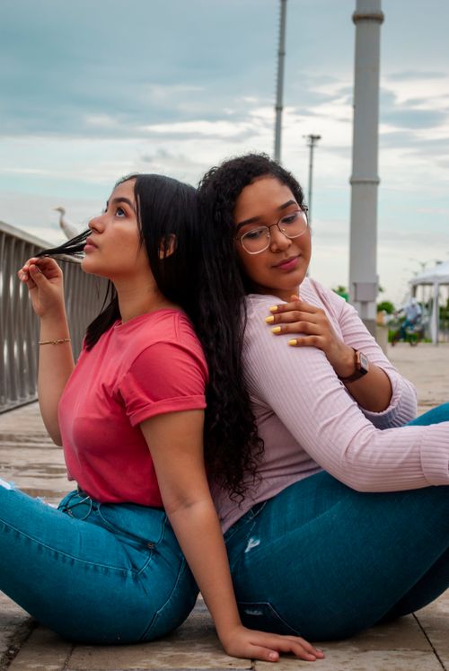Free Two women with long wavy hair sitting back to back outdoors, sharing a peaceful moment. Stock Photo