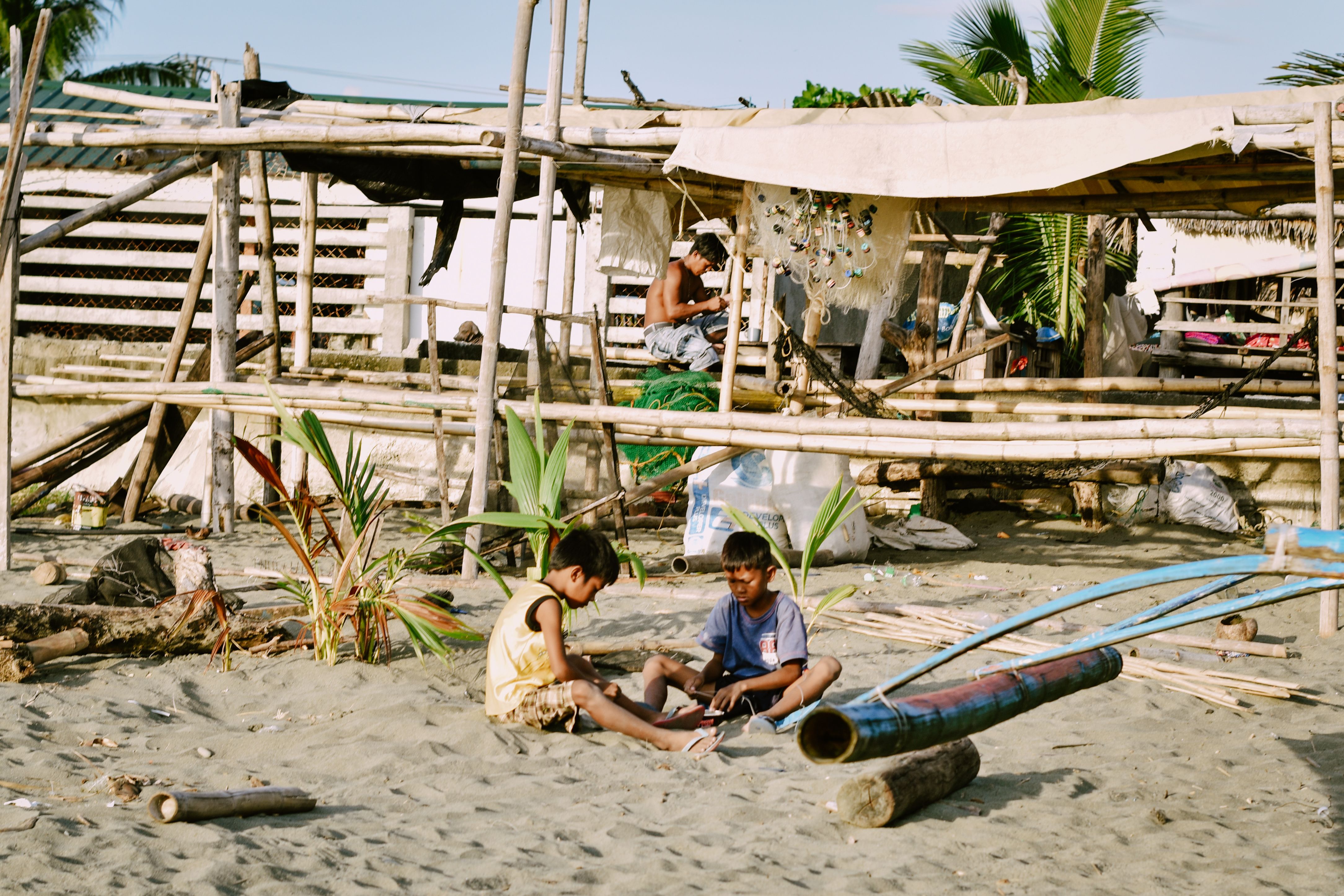Children engage in play on a sandy beach near rustic bamboo huts, under a bright sun.