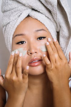 Close-up of an Asian woman applying facial cream with a towel on her head in a spa-like setting.