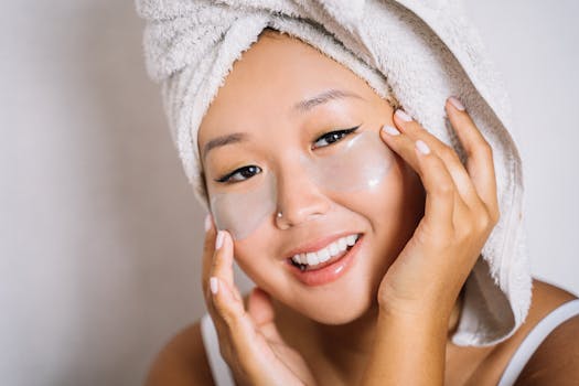 Asian woman smiling with eye patches and towel headwrap, embracing skincare.