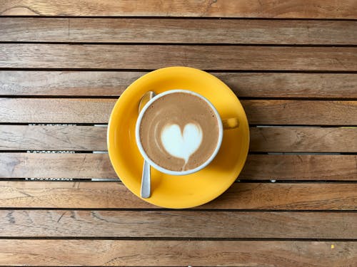 Free Top view of yellow cup of hot chocolate on saucer with heart shaped latte art placed on lumber table Stock Photo
