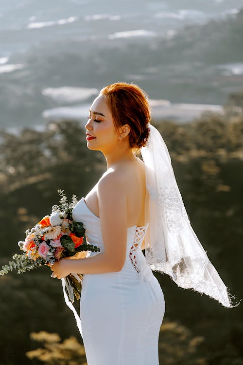 Serene young Asian female with bouquet in wedding dress