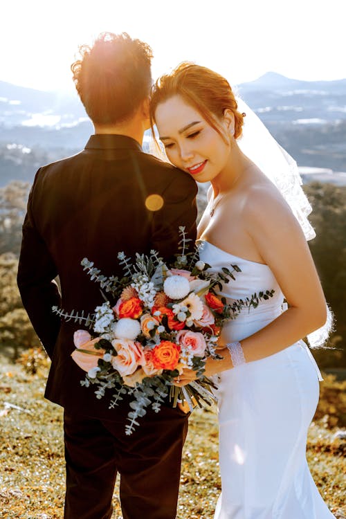 Happy Asian bride with bunch of flowers hugging groom while standing together during wedding day in green meadow